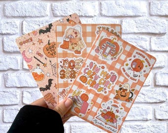 Pode incluir: Três autocolantes decorativos com tema de outono são segurados em frente a uma parede de tijolos brancos. Os autocolantes apresentam abóboras, morcegos e o texto "Spooky", "Boo to Yo", "In my cozy girl era" e "Pumpkin Season".