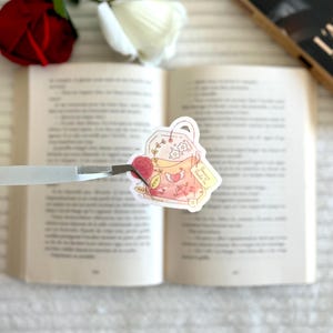Peut inclure: Un autocollant représentant un sachet de thé rose avec des motifs floraux et en forme de cœur, tenu par une pince à épiler au-dessus d'un livre ouvert. Une rose rouge et blanche est visible en arrière-plan.