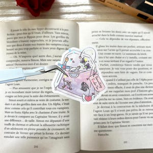 Peut inclure: Un autocollant holographique avec un motif de sachet de thé, comprenant un dragon, des cœurs et des papillons. Le texte "Romantasy Potion Tea" est visible. L'autocollant est tenu par une pince à épiler, posé sur un livre ouvert avec du texte français.