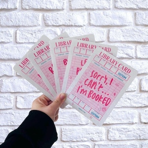 Könnte beinhalten: Eine Hand hält einen Fächer aus rosa-weißen Bibliothekskarten. Jede Karte trägt oben die Aufschrift "LIBRARY CARD", und eine Karte zeigt den Text "Sorry, I can't... I'm BOOKED". Die Karten haben ein Vintage-Design.
