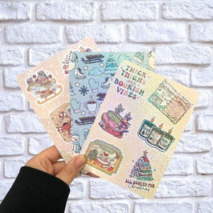 Pode incluir: Uma mão segura três autocolantes decorativos com tema natalício. Os autocolantes apresentam ilustrações de livros, um trenó, uma árvore de Natal e texto que diz "Thick Thighs and Bookish Vibes" e "All Booked for Christmas."