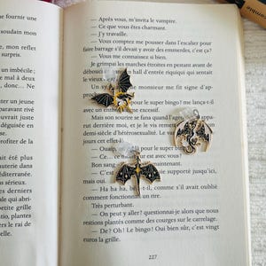 Könnte beinhalten: Drei metallene Fledermaus- und Drachenanhänger mit schwarzem Emaille und goldenen Akzenten werden auf einem offenen Buch gezeigt. Die Anhänger haben komplizierte Details und sind an durchsichtigen Ohrhaken befestigt.
