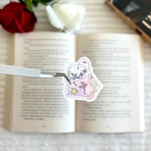 Könnte beinhalten: Ein skurriler Aufkleber in Form eines Teebeutels mit einem Fantasy-Trank-Design. Der Aufkleber zeigt eine Krone, Drachen, Schmetterlinge und eine Zaubertrankflasche. Der Text "Fantasy Potion" ist ebenfalls enthalten. Der Aufkleber wird von einer Pinzette gehalten, mit einem Buch und Rosen im Hintergrund.