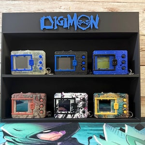 May include: A black display shelf with the word "DIGIMON" in blue. The shelf holds six Digivice toys in various colours, including blue, black, clear, and camouflage patterns. The toys have rectangular screens and button details.