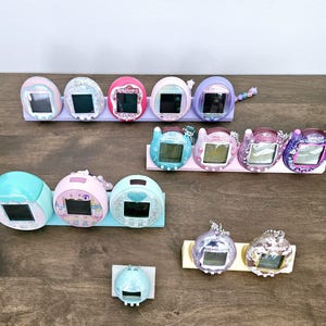May include: A collection of colorful, vintage handheld digital pets in various colors, including pink, blue, purple, and teal. Each device has a small screen and is displayed on a wooden surface.