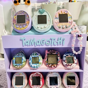 Peut inclure: Une collection de huit animaux de compagnie numériques Tamagotchi colorés disposés sur une étagère à deux niveaux. Les Tamagotchis sont disposés en rangée sur chaque étagère, la tablette supérieure présentant trois Tamagotchis roses, bleus et violets. La tablette inférieure présente cinq Tamagotchis de différentes couleurs, dont le rose, le rouge, le bleu et le violet.