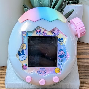 May include: A pastel-coloured digital pet device with a rainbow top and pink buttons and knob. The device features a black screen and colourful cartoon character illustrations around the screen. The device is sitting on a grey surface.