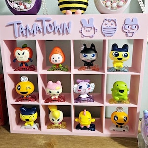 May include: A pink shelf with the words "TamaTown" at the top. The shelf has 12 compartments, each containing a different Tamagotchi character. The characters are all in various poses and colours.