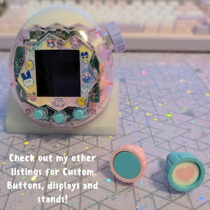 May include: A clear, pink and white handheld digital pet game with a black screen and colourful character graphics. Two custom buttons, one pink and one blue, are next to the game. The image text says, "Check out my other listings for Custom Buttons, displays and stands!"