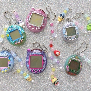 May include: A collection of six Tamagotchi digital pet devices in various colours and patterns, including pink glitter, blue floral, and purple butterfly designs. Each device features a screen, buttons, and a beaded chain with small charms.