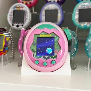May include: A pink Tamagotchi digital pet with a starry night display. The device has a pink frame with green buttons and a green top. Other Tamagotchi devices in various colours are visible in the background.
