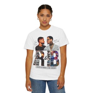 K&ouml;nnte beinhalten: Wei&szlig;es T-Shirt mit einer Grafik von Usher Raymond und Chris Brown. Das Design enth&auml;lt den Text "RAYMOND & BROWN" und die Namen der K&uuml;nstler. Das Shirt hat einen Rundhalsausschnitt und kurze &Auml;rmel.
