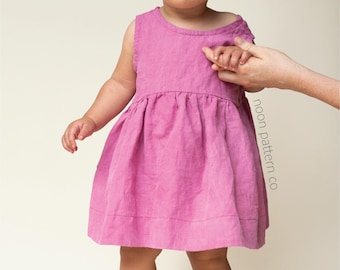 May include: A sleeveless, pink dress with a gathered skirt on a young child. The dress is made of a soft fabric and has a simple, round neckline. The child is standing and being held by an adult's hand. The text "noon pattern co" is visible.
