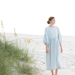 Puede incluir: Vestido de lino azul claro con mangas abullonadas de tres cuartos. El vestido tiene una cintura fruncida y una falda fluida. La modelo está de pie en una playa de arena con hierba alta en primer plano.
