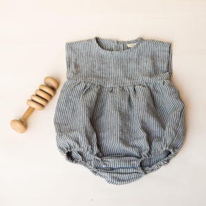May include: A blue and white striped baby romper with a round neckline and gathered waist. A wooden rattle is next to the romper. The romper has a button closure at the back.