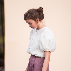 May include: A white linen blouse with short, puffed sleeves and a round neckline. The blouse is buttoned down the front with small, round buttons. The model is wearing a pair of mauve-colored pants. The background is a solid, light peach color.