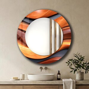 May include: A round decorative mirror with a unique design. The frame features a layered pattern in shades of orange, brown, and gray. The mirror is mounted above a wooden vanity with a white sink and a small potted plant.
