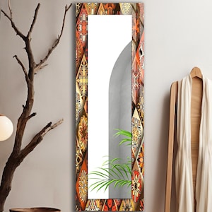 Pode incluir: Um espelho de parede alto e retangular com uma moldura decorativa apresentando um padrão repetitivo de motivos geométricos e florais em tons de laranja, amarelo e castanho. A superfície reflete uma planta verde e uma parede cinzenta curva.