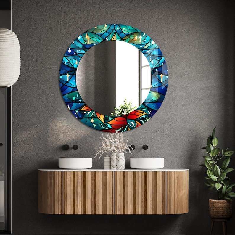 May include: A round mirror with a blue, green, and orange stained glass design. The mirror is hanging on a gray wall above a wooden vanity with two sinks.