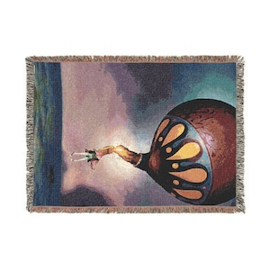May include: Woven throw blanket with a surrealist design. The artwork features a large, ornate object with a butterfly-like pattern in shades of brown, blue, and gold. The background has a cloudy, atmospheric effect. The blanket has a fringed edge.