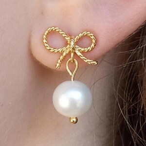 May include: A gold-colored bow-shaped earring with a rope-like texture, attached to a large, white, round pearl. The earring is worn on the ear. The pearl has a small gold-colored bead at the bottom.