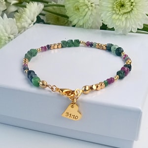 May include: A beaded bracelet featuring green and maroon gemstones interspersed with gold-colored beads. The bracelet has a gold-colored clasp and a heart-shaped charm with the inscription "31:10". The bracelet is displayed on a white surface.