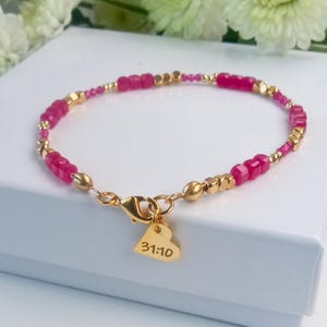May include: A delicate bracelet featuring a combination of gold-colored beads and vibrant pink gemstone beads. A small gold heart charm with the inscription "31:10" dangles from the clasp. The bracelet is presented on a white surface, suggesting a jewelry theme.