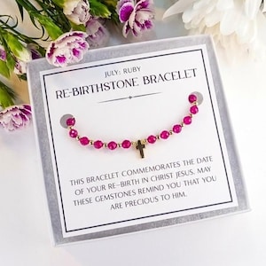 May include: A ruby birthstone bracelet with a gold cross charm, presented in a gift box. The card reads "JULY: RUBY RE-BIRTHSTONE BRACELET". The bracelet is adorned with red gemstones and gold beads.