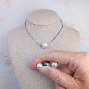 May include: A silver-toned chain necklace featuring a single cream-colored pearl pendant. Three additional loose pearls in light blue, dark grey, and light pink are shown separately, each with a visible hole for threading onto a chain.