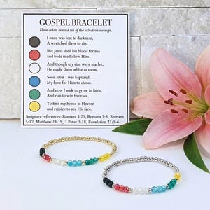 May include: Two beaded Gospel bracelets with gold and silver accents, each featuring a sequence of black, red, white, blue, green, and yellow beads. A card with text about the salvation message is in the background, along with a pink flower.