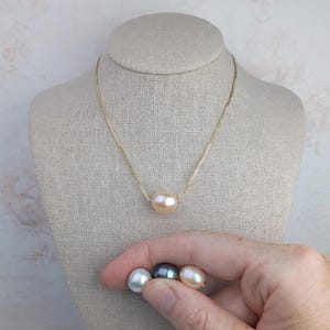 May include: A gold-toned chain necklace featuring a single, centered, round, light peach-colored pearl. Below the necklace, three additional loose pearls are shown in white, dark grey, and light peach, each with a smooth, lustrous finish.