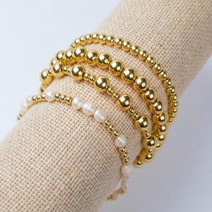 May include: Four gold beaded bracelets and one pearl bracelet are displayed on a beige fabric roll. The gold bracelets have varying bead sizes, while the pearl bracelet features small gold beads interspersed with white pearls.
