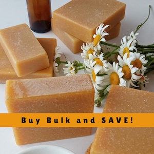 May include: Several rectangular bars of light brown soap are arranged with a small brown bottle and a bouquet of white daisies with yellow centers. The text "Buy Bulk and SAVE!" is displayed on an orange banner.