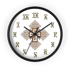 Op de afbeelding: Een ronde wandklok met een zwarte rand en een witte wijzerplaat. Het midden toont een gedetailleerd bronskleurig Ethiopisch kruis. Twaalf donker olijfgroene Ge'ez-tekens zijn in een cirkel rondom het centrale kruismotief geplaatst.