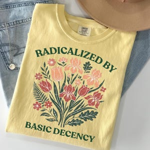 May include: A pale yellow t-shirt featuring a central graphic of a floral bouquet with red, pink, and orange flowers and green leaves. The text RADICALIZED BY is printed in dark green above the flowers, and BASIC DECENCY is printed below.