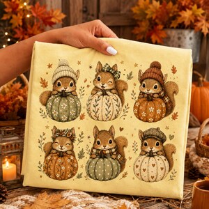 May include: A light yellow fabric featuring a repeating pattern of six illustrated squirrels, each sitting behind a different colored pumpkin. The squirrels wear various accessories like hats, scarves, and floral crowns, surrounded by small autumn leaves.