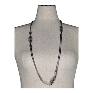 May include: A long, dark silver necklace with ornate, filigree-style pendants. The necklace is displayed on a mannequin and features multiple strands of delicate chain. The pendants are symmetrical and add a vintage or antique aesthetic.