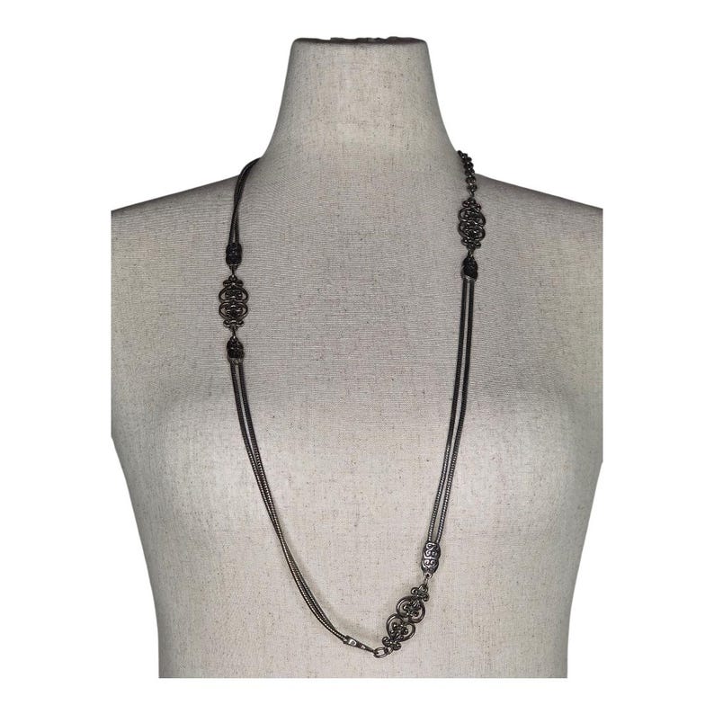 May include: A long, dark silver necklace with ornate, filigree-style pendants. The necklace is displayed on a mannequin and features multiple strands of delicate chain. The pendants are symmetrical and add a vintage or antique aesthetic.