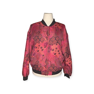 May include: A vibrant pink bomber jacket with a floral pattern in shades of red and black. The jacket features a black collar, cuffs, and waistband, and a full-length zipper. The jacket is displayed on a mannequin.
