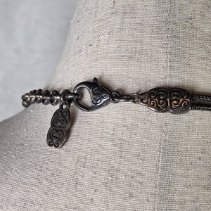 May include: Close-up of a silver-toned necklace clasp and chain. The necklace features an ornate clasp with a decorative design and a small pendant. The chain is composed of small, linked beads.