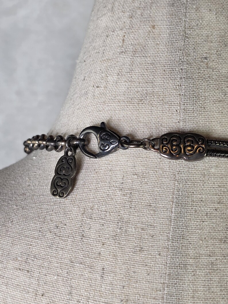 May include: Close-up of a silver-toned necklace clasp and chain. The necklace features an ornate clasp with a decorative design and a small pendant. The chain is composed of small, linked beads.