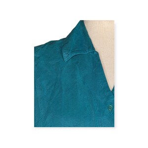May include: Teal-colored collared shirt. The top of the shirt is visible, showing the collar and a portion of the buttoned front. The fabric has a slightly textured appearance. A single green button is visible.