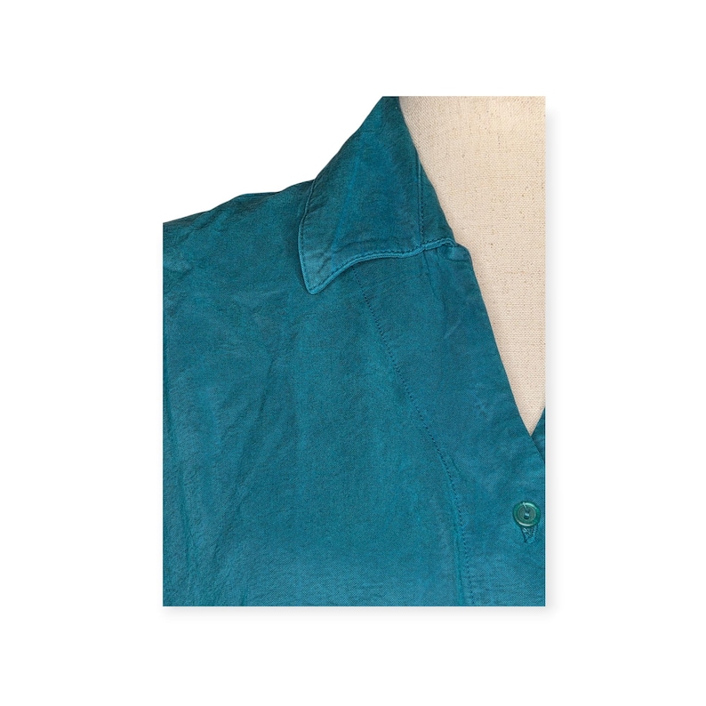 May include: Teal-colored collared shirt. The top of the shirt is visible, showing the collar and a portion of the buttoned front. The fabric has a slightly textured appearance. A single green button is visible.