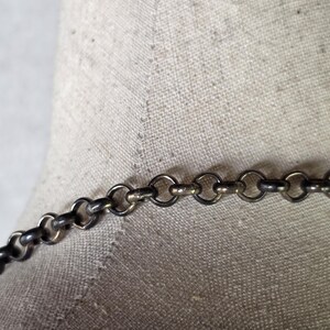 May include: A close-up shot of a silver-coloured chain necklace featuring small, circular links. The necklace is set against a neutral fabric backdrop, emphasising its metallic lustre and design. The jewellery appears to be a delicate piece.
