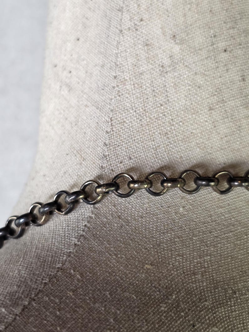May include: A close-up shot of a silver-coloured chain necklace featuring small, circular links. The necklace is set against a neutral fabric backdrop, emphasising its metallic lustre and design. The jewellery appears to be a delicate piece.