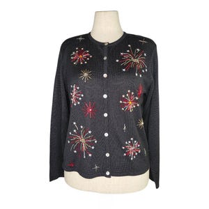Könnte beinhalten: Schwarze Strickjacke mit gestickten Feuerwerkskörpern in Rot, Gold und Weiß. Der Pullover hat eine Knopfleiste vorne und das Jahr "2000" auf der Vorderseite gestickt. Der langärmlige Pullover wird auf einer Schaufenquin präsentiert.