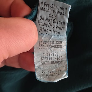 May include: A teal fabric tag with care instructions and product information. The tag reads "Pre-Shrunk", "Machine-Wash Cold", "Do Not Bleach", "Tumble Dry Warm", and "Steam Iron". The tag also includes the website TIANELLO.COM, a phone number, and the style name "SUZY BLOUSE".