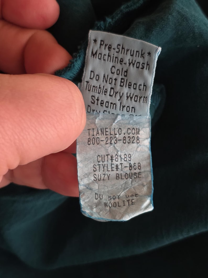 May include: A teal fabric tag with care instructions and product information. The tag reads "Pre-Shrunk", "Machine-Wash Cold", "Do Not Bleach", "Tumble Dry Warm", and "Steam Iron". The tag also includes the website TIANELLO.COM, a phone number, and the style name "SUZY BLOUSE".