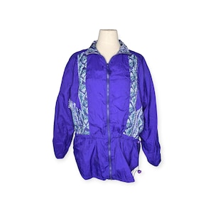 Peut inclure: Veste de survêtement violette vintage avec fermeture éclair complète. Le devant et les épaules présentent un motif à motifs turquoise et blanc. La veste a une taille à cordon et des manches longues.