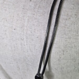 May include: Close-up of a black metal necklace with a snake chain design. The necklace has a detailed pendant and a textured clasp. The background is a light-coloured fabric.